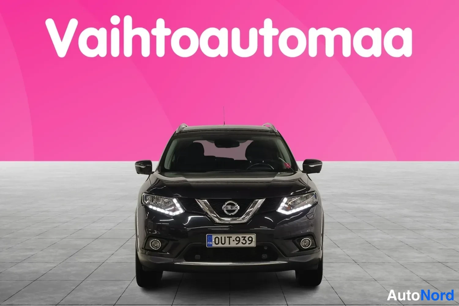 Edestä: Musta auto suoraan edestäpäin kuvattuna, rekisterikilpi, päiväajovalot päällä.