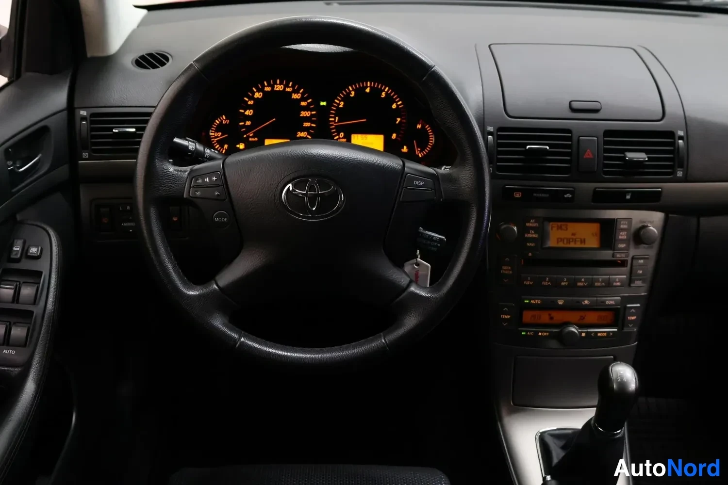 Muu: Auton sisätilan kojelauta ja Toyota-ohjauspyörä, mittaristo valaistu, manuaalivaihteen keila ja radio näkyvissä.