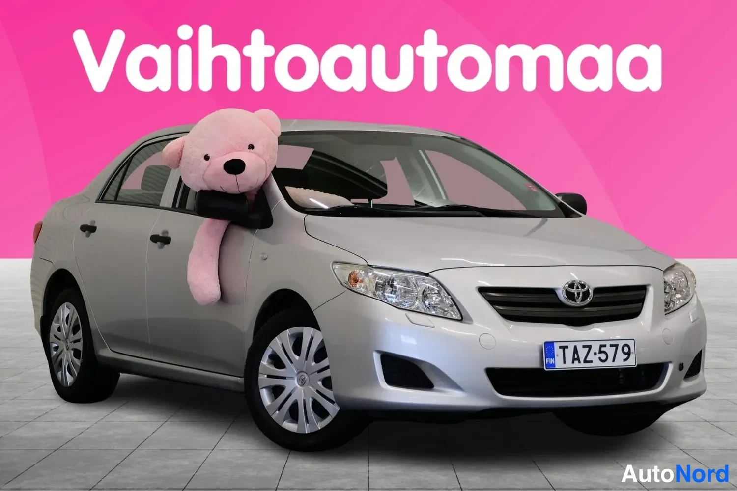 Etuviisto, nokka oikealle: Hopeanvärinen Toyota Corolla -henkilöauto kuvattu kolmannelta osapuolelta edestä oikealta, keulakilpi, pinkki tausta.