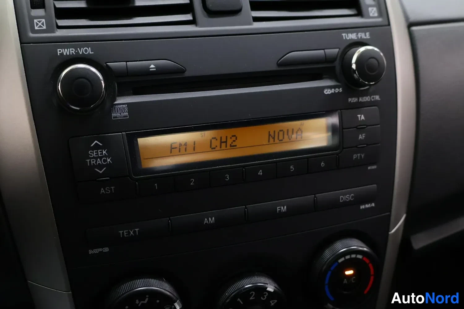 Yksityiskohta: Keskikonsolin radiopaneeli lähikuva; näytöllä teksti 'FM1 CH2 NOVA', painikkeet TEXT, AM, FM, DISC, A/C-säädin ja nuppia 1-4.