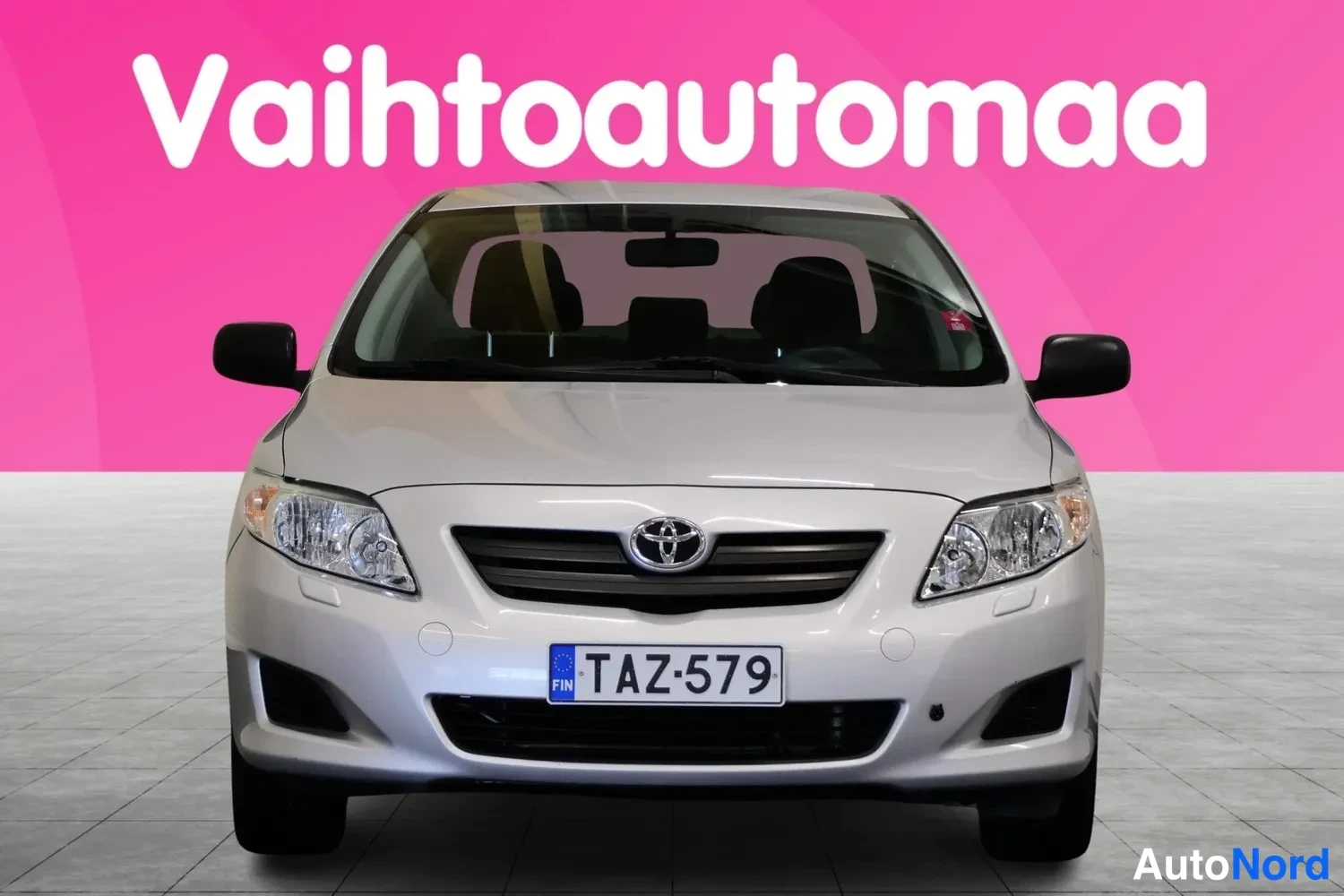 Edestä: Hopeanvärinen Toyota Corolla -henkilöauto suoraan edestä kuvattuna, keulakilpi, pinkki tausta.