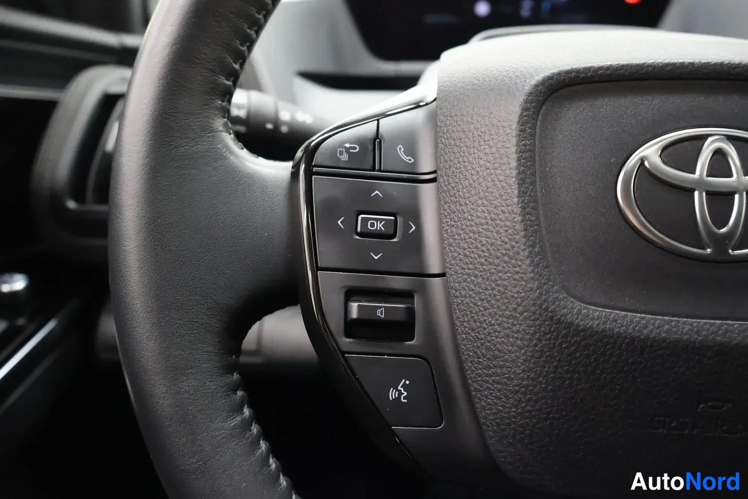 Muu: Lähikuva ohjauspyörän vasemmasta puhelin- ja ääniohjainnäppäimistä, joissa näkyy puhelin-, OK-, äänenvoimakkuus- ja ääniohjausmerkit, Toyota-merkki oikealla.
