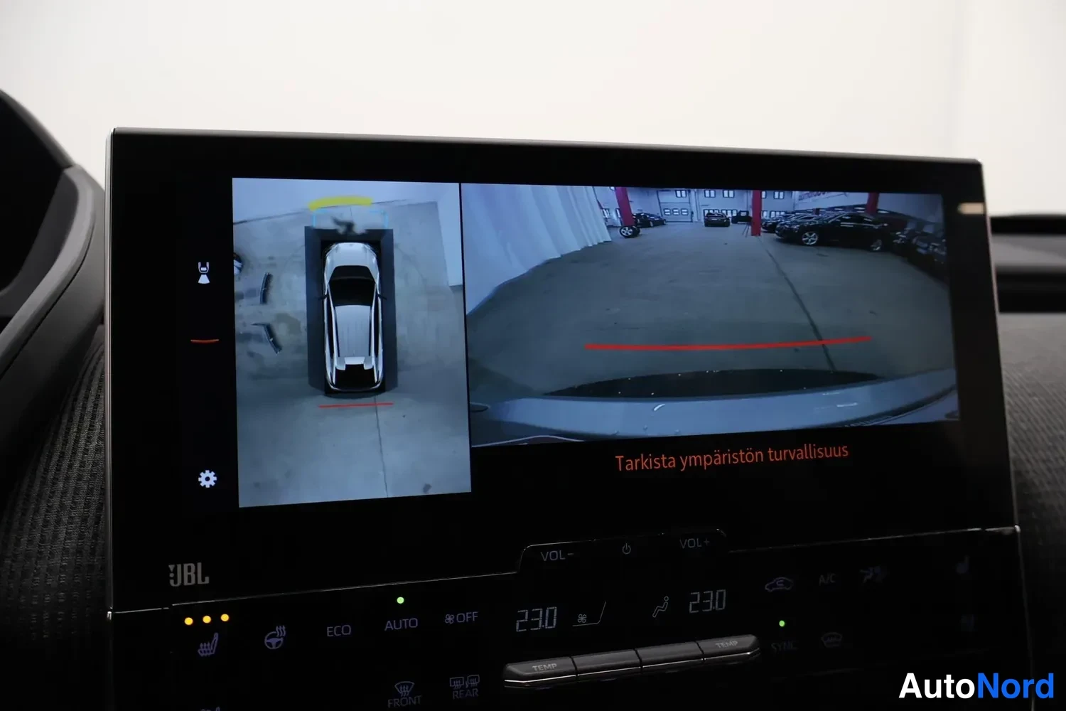 Yksityiskohta: Keskikäytävän suuri kosketusnäyttö näyttää 360-asteen kamerakuvan autosta ylhäältä ja takakameran näkymän, tekstillä 'Tarkista ympäristön turvallisuus'; näytön
