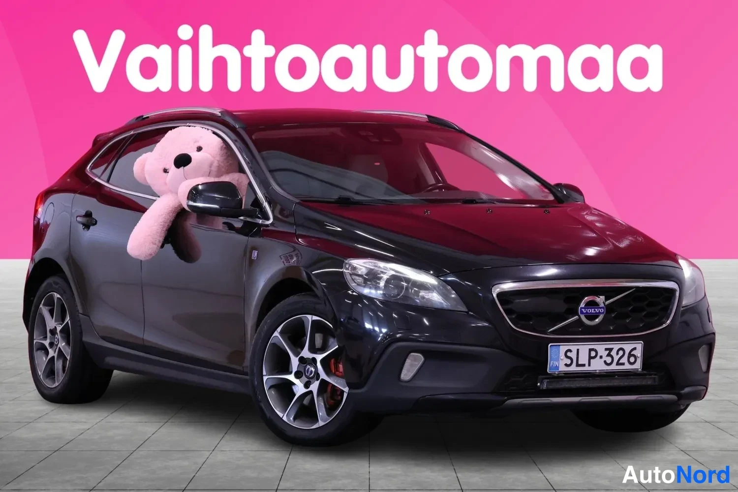 Etuviisto, nokka oikealle: Musta Volvo etuviistosta oikealta: keulailme, rekisterikilpi, kattokaiteet ja kevytmetallivanteet; etuikkunassa vaaleanpunainen pehmolelu.