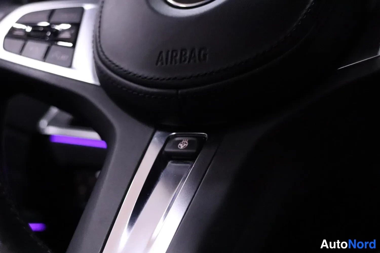 Muu: Lähikuva ohjauspyörän keskiosasta: AIRBAG-teksti ilmatyynyn ovella, ohjauspyöränlämmityskuvake metallipinnoitteisessa painikkeessa ja ohjauspyörän puhallinpaini