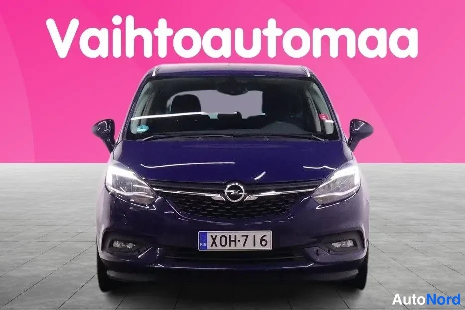 Edestä: Tummansininen tila-auto suoraan edestä kuvattuna, rekisterikilpi, Opel-merkki etusäleikössä, vaaleanpunainen tausta.