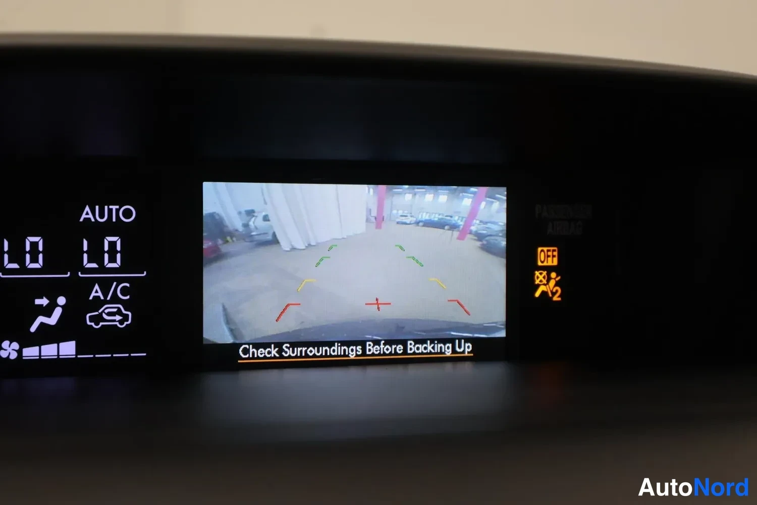 Yksityiskohta: Kojelaudan näyttö, jolla näkyy peruutuskameran kuva huoltosalista tekstillä Check Surroundings Before Backing Up.