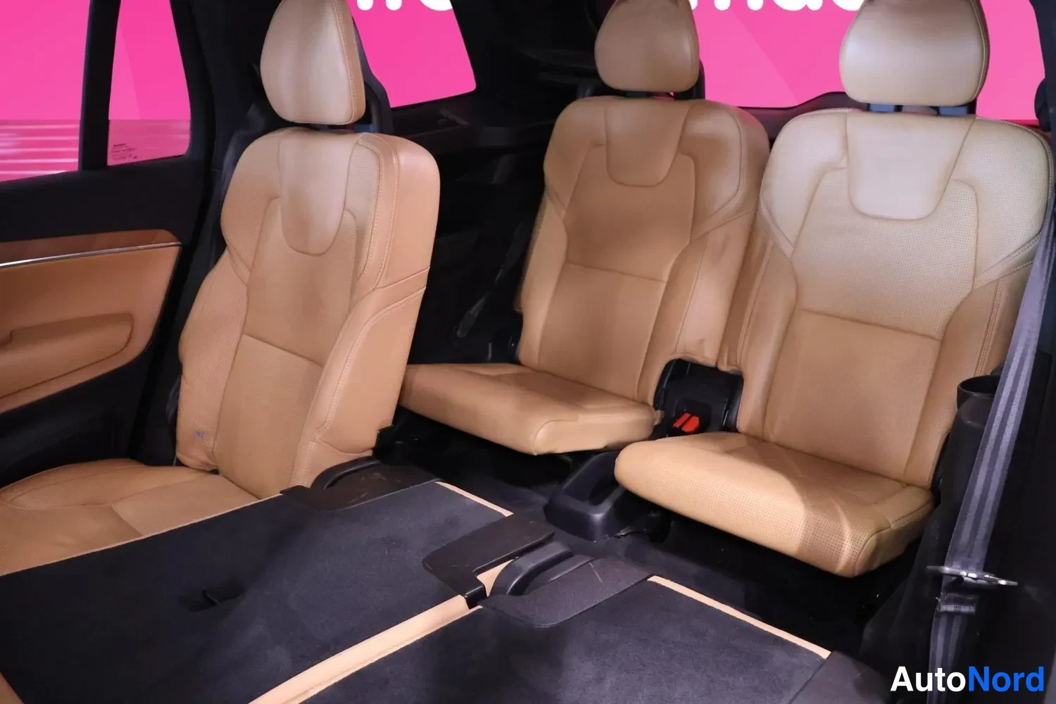 Takaistuimet: Tämän kuvan sisäkulmasta nähdään kolme takapenkkiä, jotka ovat ruskeaa nahkaa. Penkit sijaitsevat autoon, jossa on tumma lattia ja punainen seinä taustalla.
