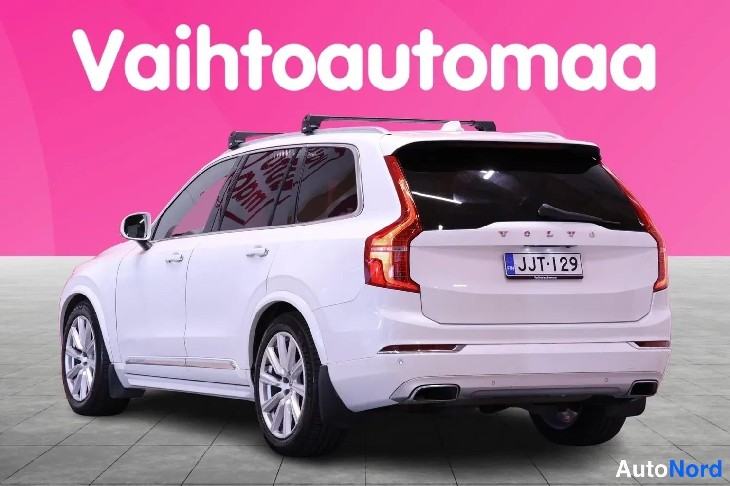 Takaviisto: Valkoinen farmari-avolava? Ei – valkoinen Volvo XC90 -tyyppinen SUV takaa kolmannevasemmalta kuvattuna; rekisterikilpi ja kattotelineet näkyvät.