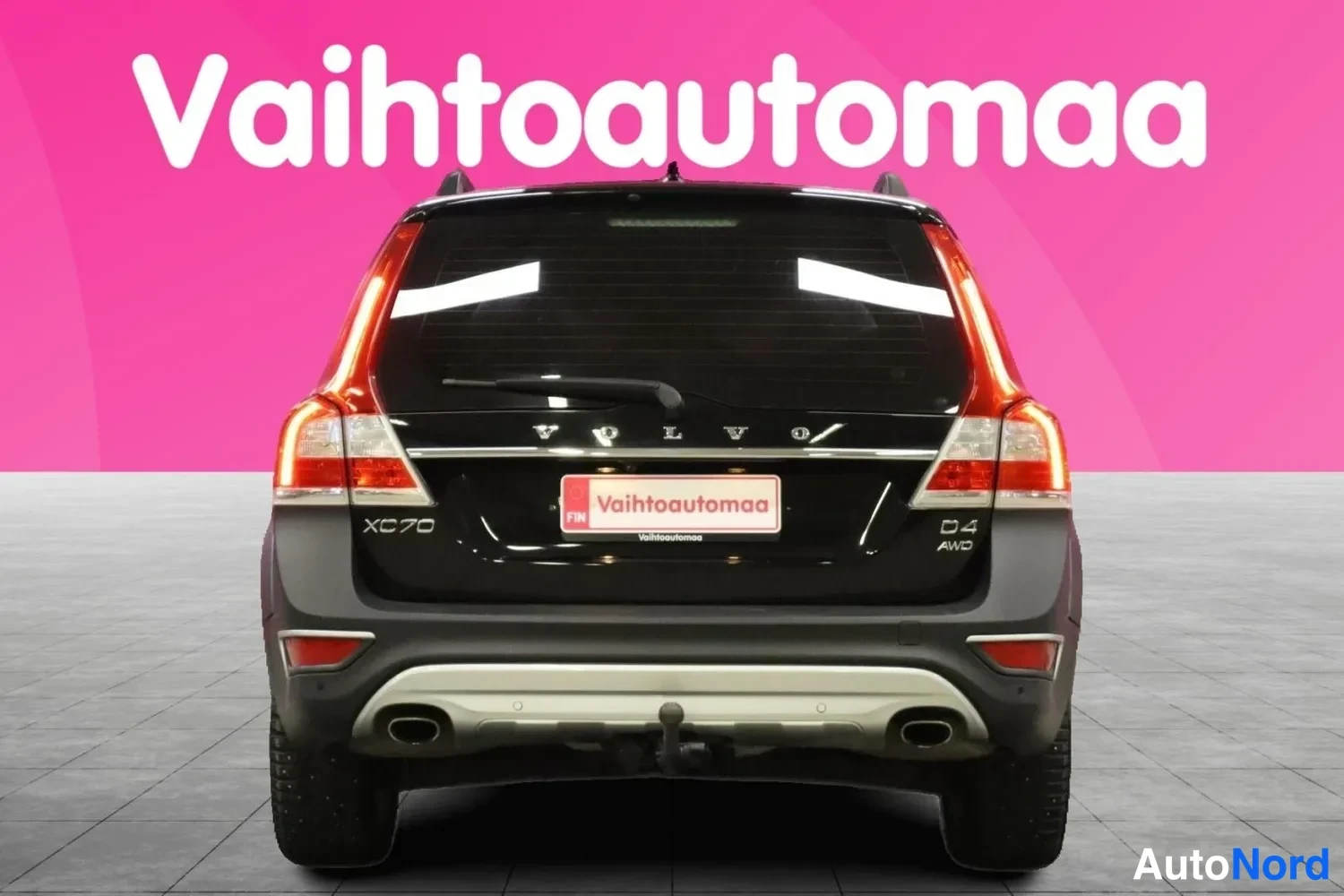 Takaa: Vaihtoautomaan markkinointikuva: musta farmariauto suoraan takaa, XC70- ja D4 AWD -merkit takaluukussa vaaleanpunaista taustaa vasten.