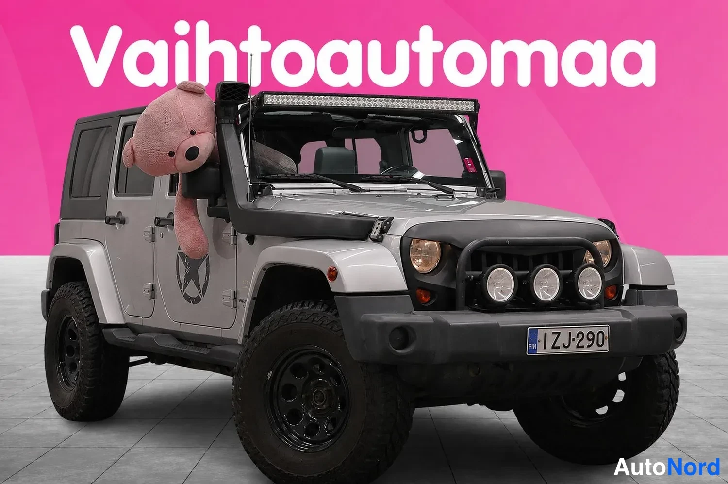 Etuviisto, nokka oikealle: Hopeanharmaa Jeep-etukolmivartalokuva, maskissa kolme lisävaloa, kattotankona LED-palkki ja ovenkolkassa suuri nallekarhu.