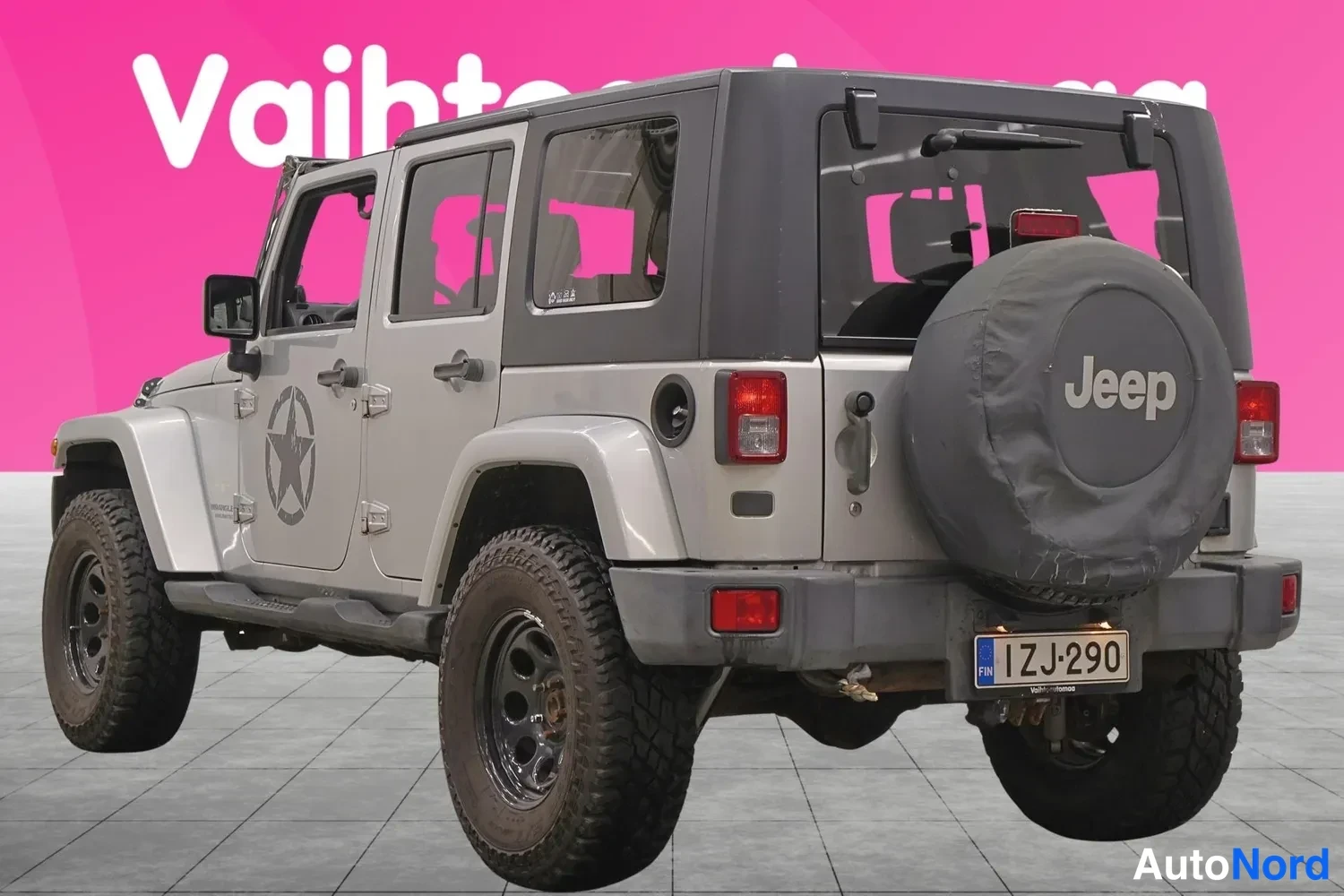 Takaviisto: Hopeanharmaa Jeep Wrangler takaa ja vasemmalta sivulta kuvattuna; pehmeä varalippu Jeep-tekstillä, rekisteritunnus, tumma kova katto ja neljä ovea näkyvissä.