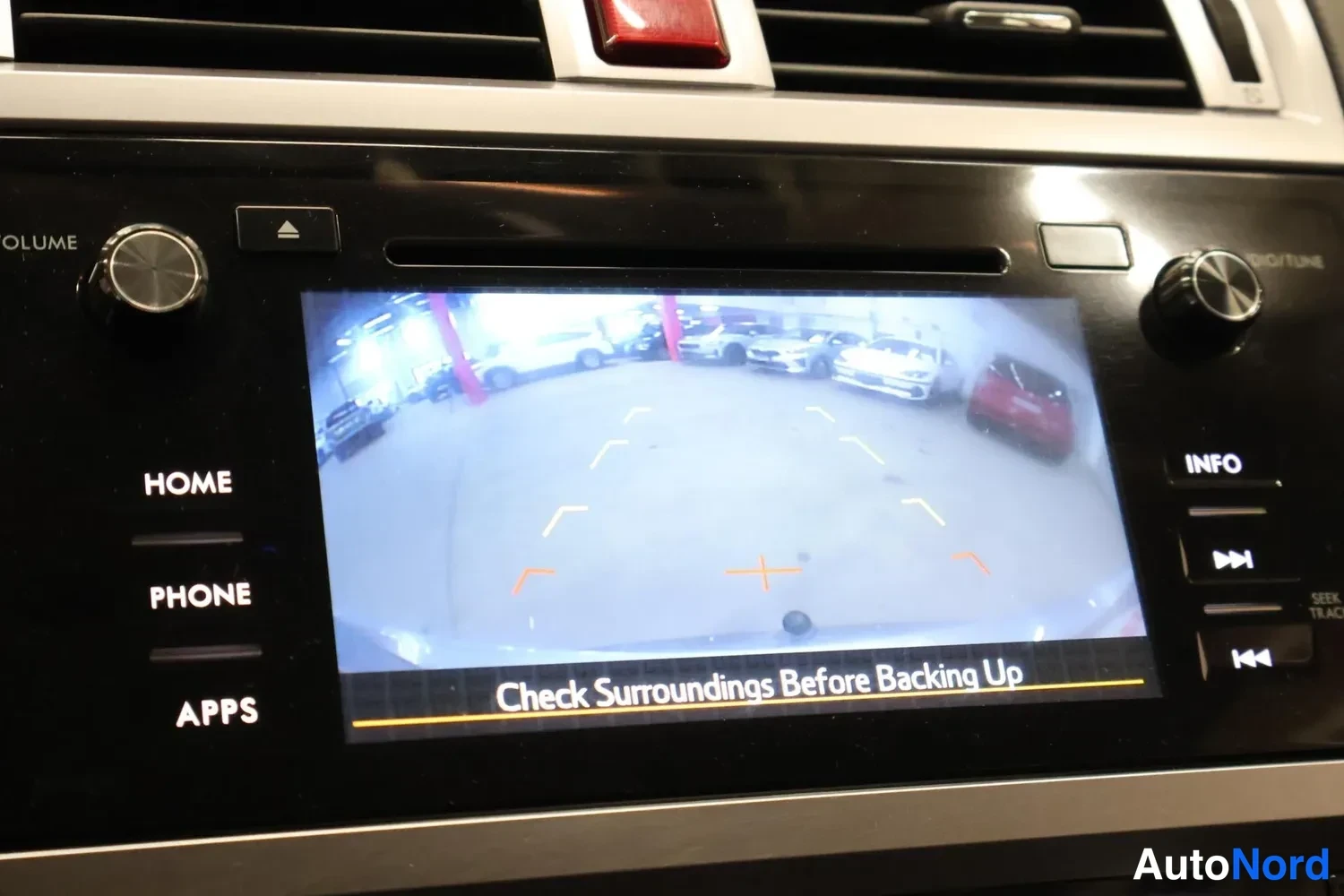 Muu: Keskikonsolin näyttö näyttää peruutuskameran kuvan tekstillä 'Check Surroundings Before Backing Up'. Reunalla HOME-, PHONE-, APPS-, INFO-painikkeet ja volume/tu