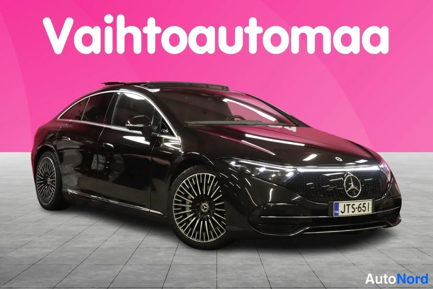 Etuviisto, nokka oikealle: Mustan Mercedes-Benz EQS:n etunurkkakuva oikealta, rekisteri, taustana pinkki vaaka ja lattia.