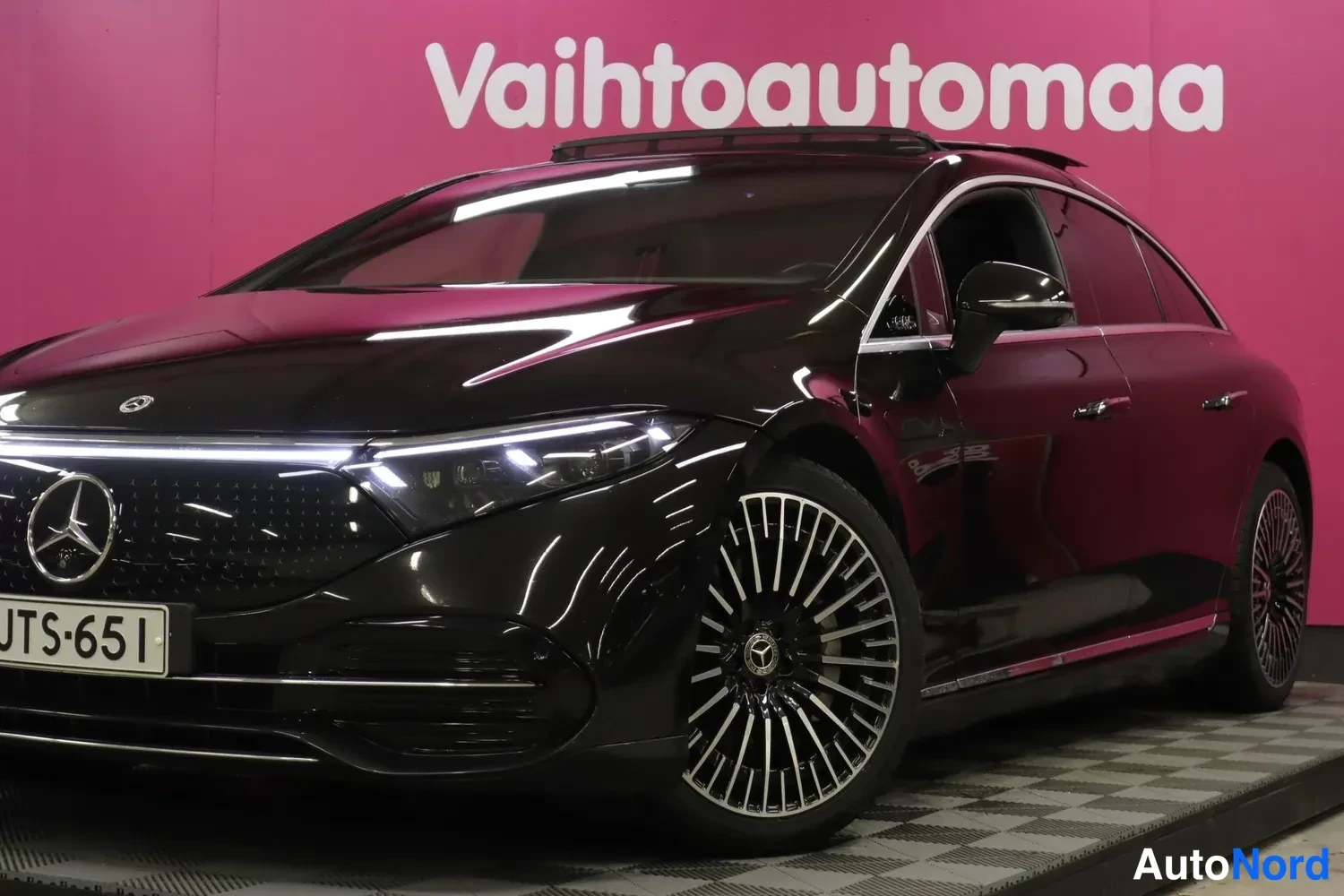 Etuviisto, nokka vasemmalle: Mustan Mercedes-Benzin etukulmanäkymä valaistulla etusäleiköllä ja monihaaraisella kevytmetallivanteella, rekisteritunnus.
