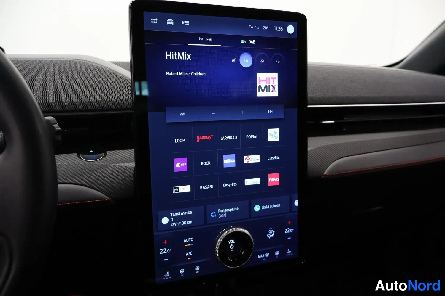 Yksityiskohta: Pystysuora infotainment-näyttö näyttää FM/DAB-radiolähteitä (HitMix, Robert Miles - Children), kanavaruudukon ja ilmastointiasetukset 22 asteen lämpötilalla.