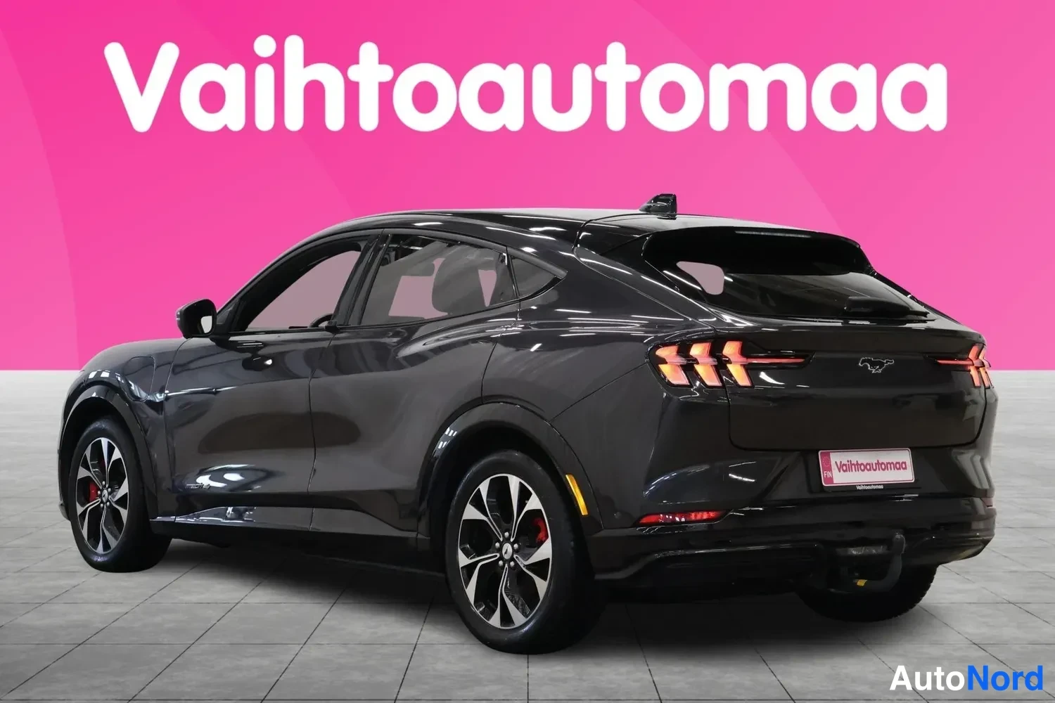 Takaviisto: Mustanharmaan tila-auton takakulmanäkymä vasemmalta, rekkiteline ja Vaihtoautomaa-kyltti taustalla.