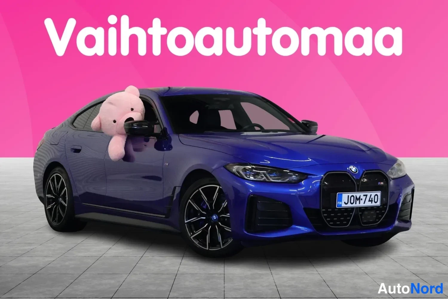 Etuviisto, nokka oikealle: Sininen BMW Coupe etuviistosta vasemmalta kuvattuna studioitaustaa vasten; pinkki pehmolelu takanäytössä, tunnus.