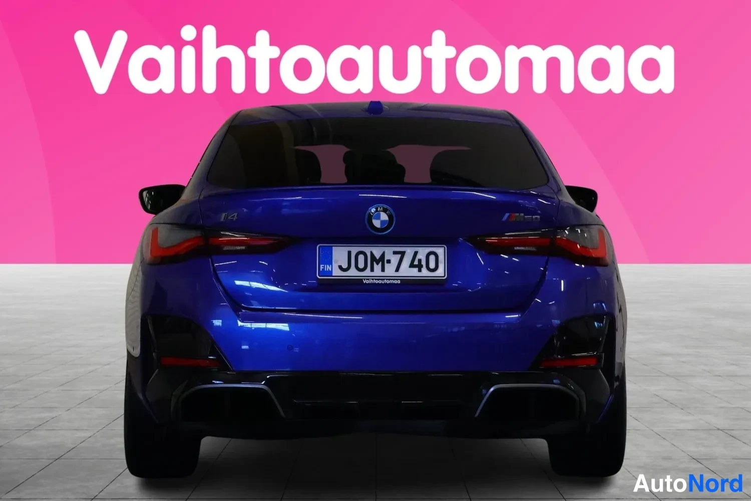 Takaa: Sininen BMW i4:n takanäkymä rekisteritunnuksella, vaaleaa lattiaa ja vaaleanpunaista taustaa vasten.