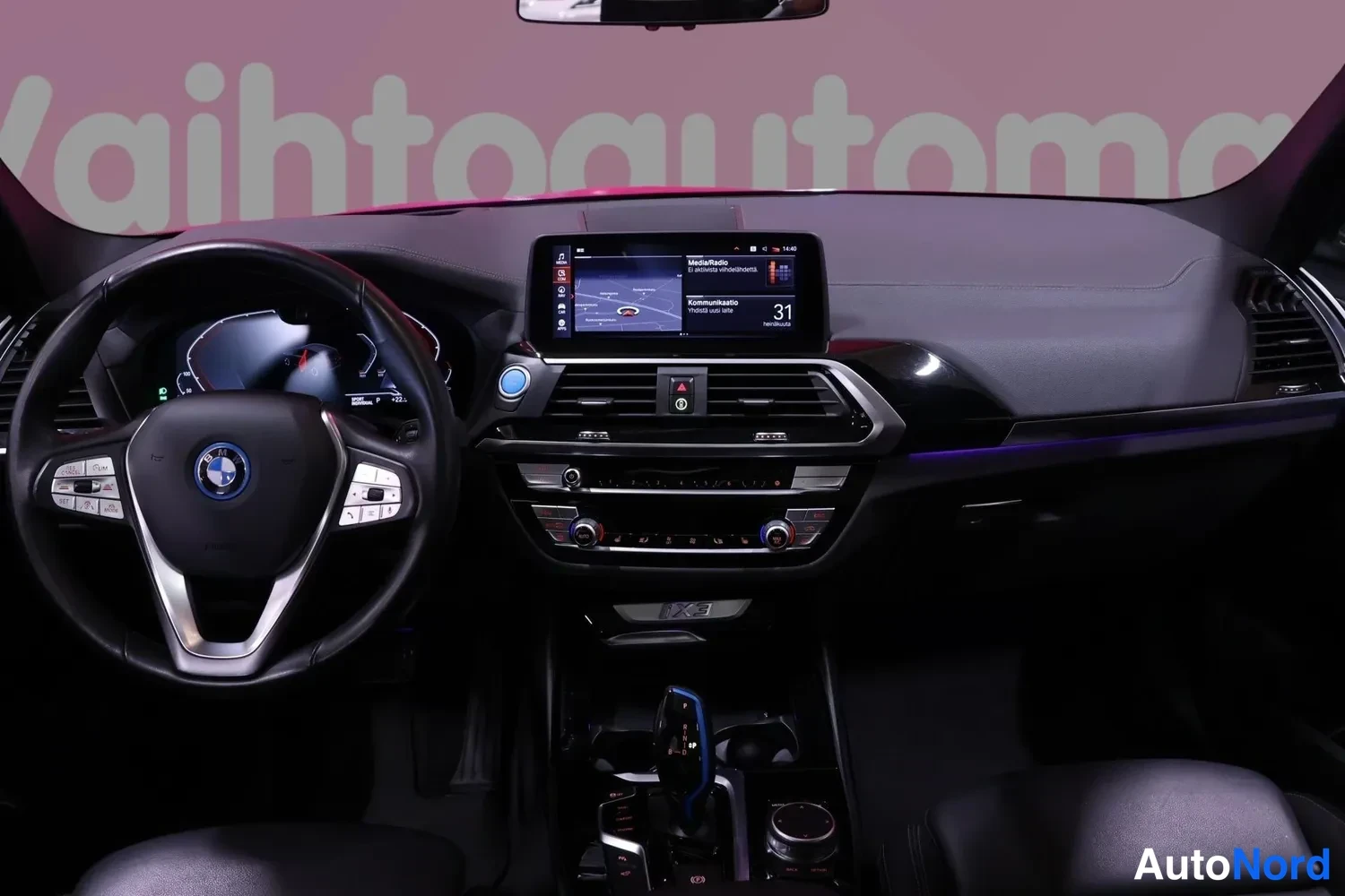 Edestä: BMW iX3:n hallintapaneeli: ohjauspyörä, digitaalinen mittaristo ja keskinäyttö, jonka kartta ja Medi&aRadio-valikko näkyvät.