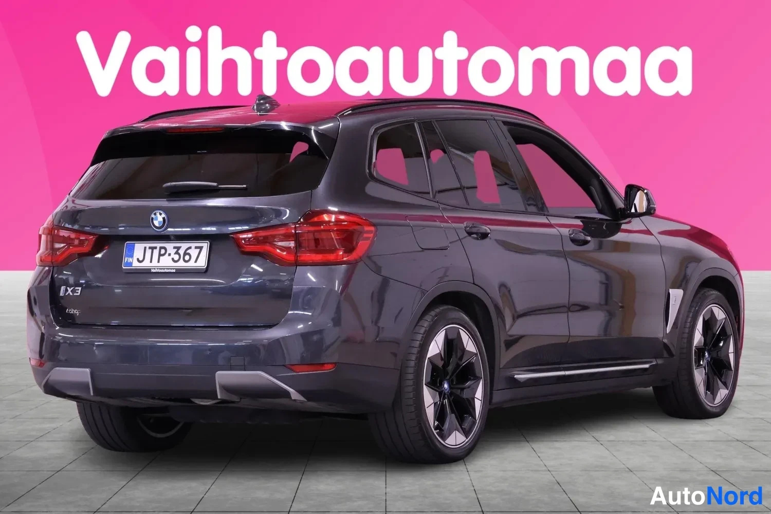 Takaviisto: Takana oikealla kuvattu tummanharmaa BMW iX3 -auto. Takavalot, rekisterikilpi ja mallinumero 'iX3' takaluukussa ovat selkeästi erottuvia. Piilotetut taka-valopa