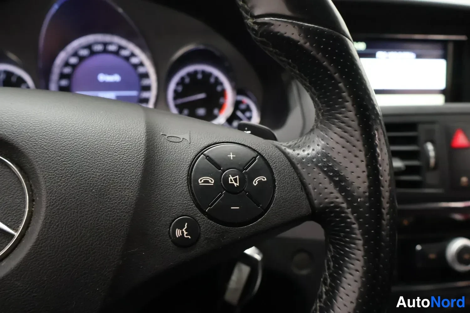 Muu: Sisätilakuva Mercedes-Benzin ratin vasemmasta puhelin- ja äänentoistonäppäimistöstä, mittaristo taustalla.