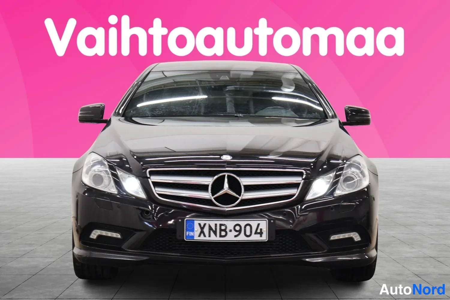 Edestä: Mustan Mercedes-Benzin etunäkymä, edessä Mercedes-tähti ja rekisterikilpi.