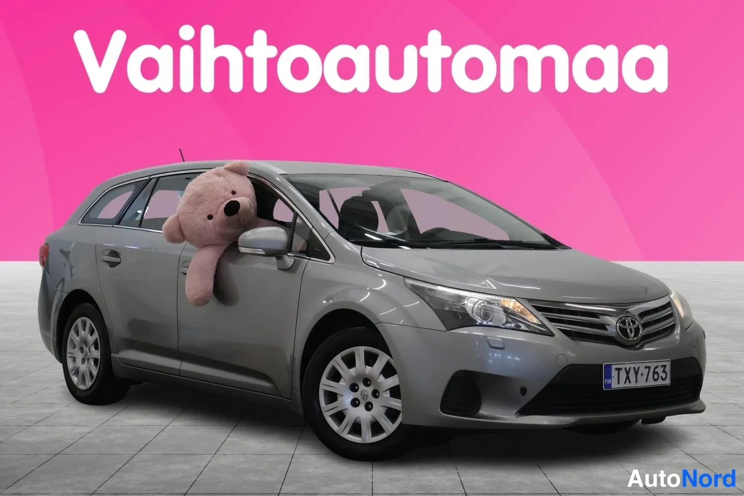 Etuviisto, nokka oikealle: Toyota Avensis -farmari kolmivärikuvassa edestä oikealta, pinkki tausta ja nalle ikkunassa.