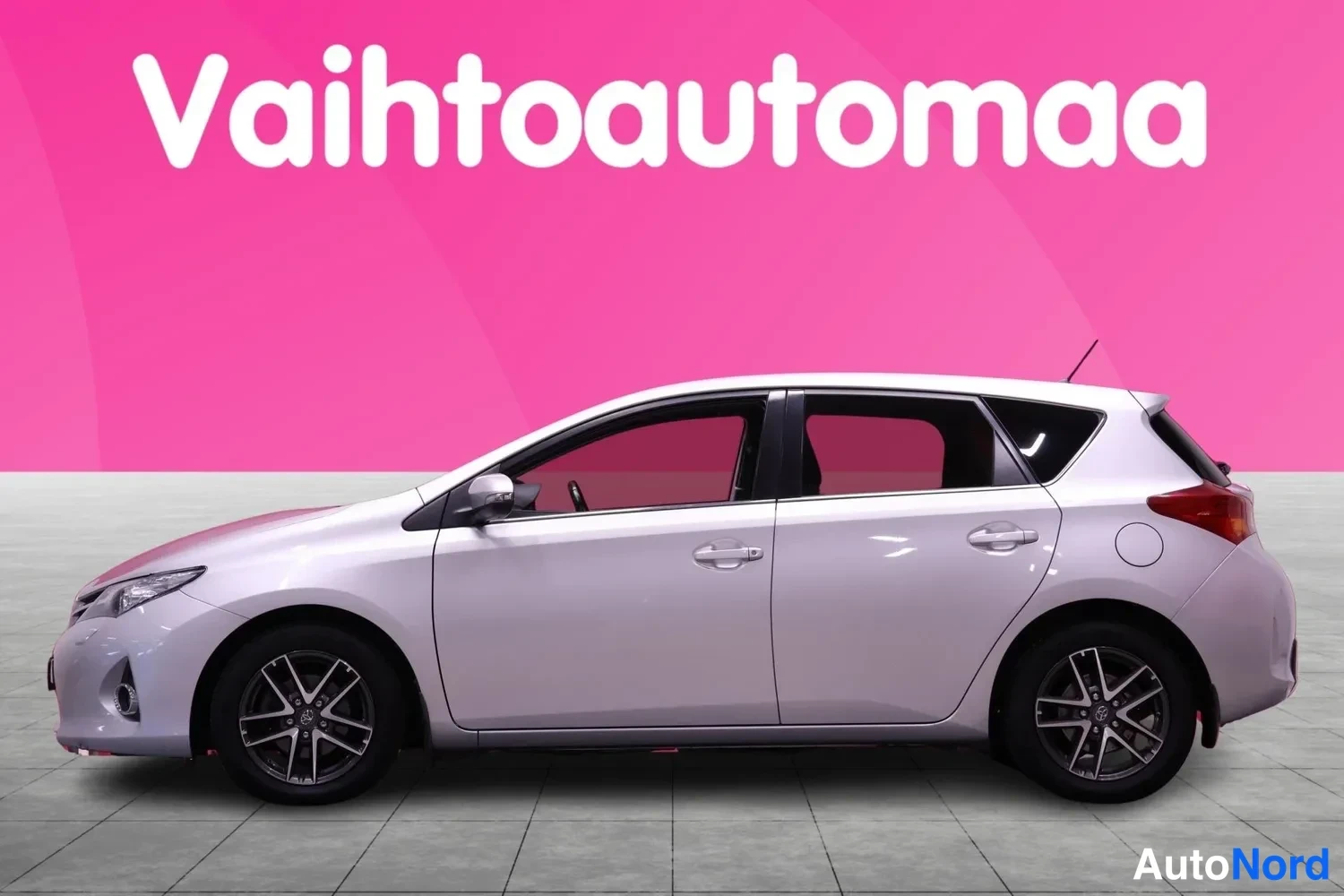 Sivulta: Auton vasen kylki sivusuunnasta, koko auto näkyvissä vaalealla lattialla ja vaaleanpunaisella taustalla.