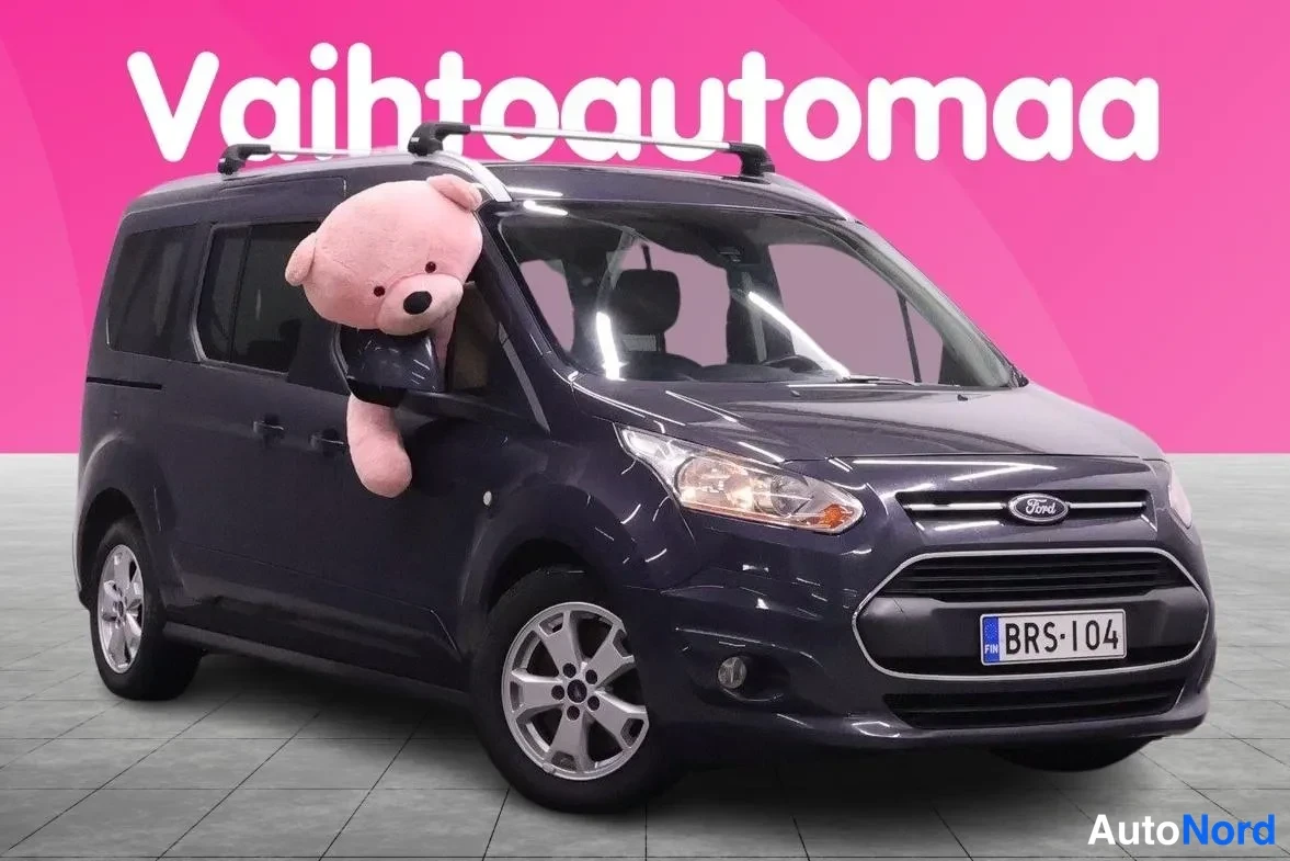 Etuviisto, nokka oikealle: Tummanharmaa Ford Tourneo etukulmasta oikealta, rekisterikilpi, kattokaiteet ja suuri pehmolelu ikkunassa.
