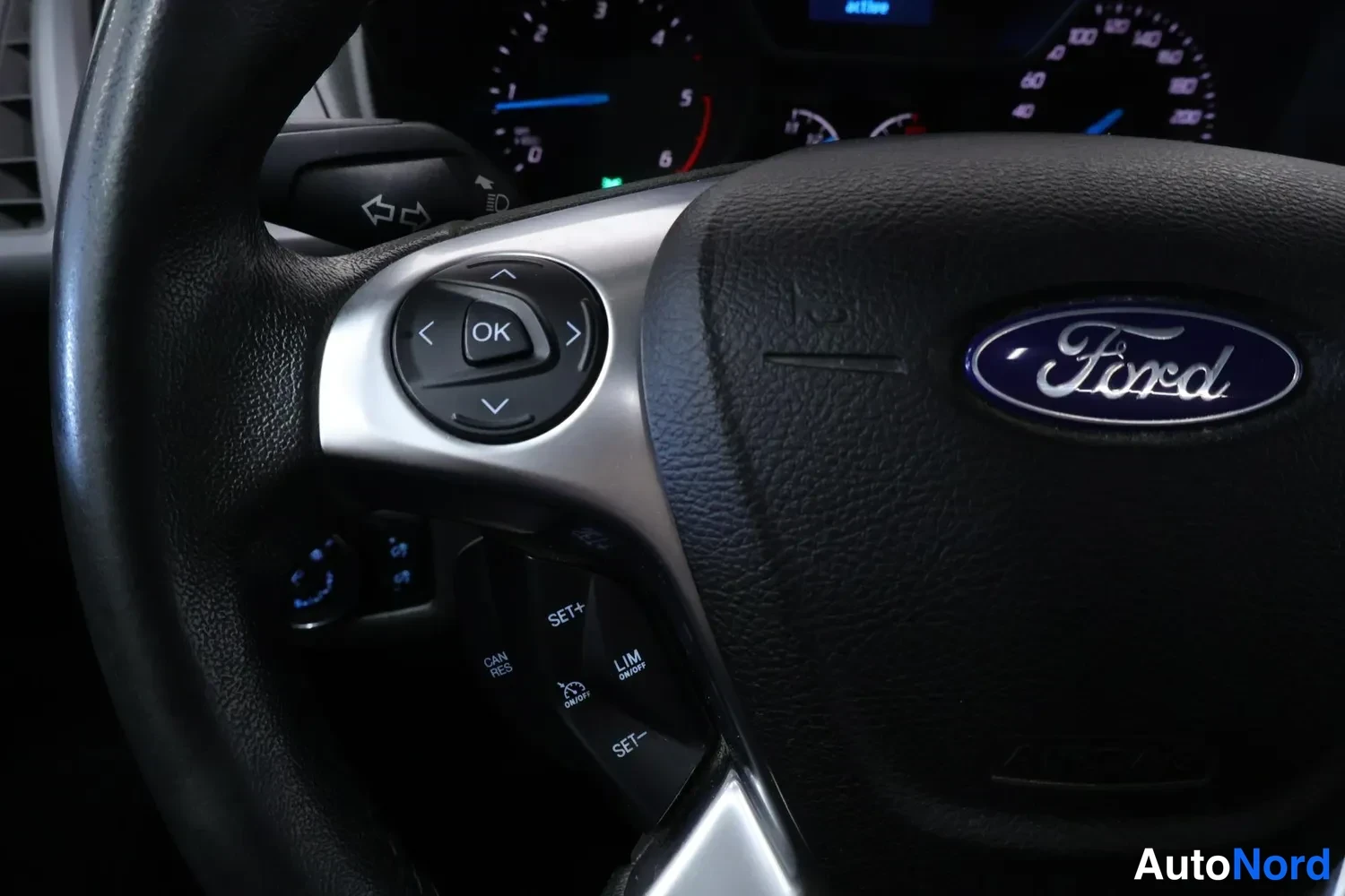 Edestä: Tarkka lähikuva ohjauspyörän vasemmasta puhallinpainikkeesta ja Ford-merkistä; painikkeissa OK, SET+, SET-, CAN RES ja LIM ON/OFF.