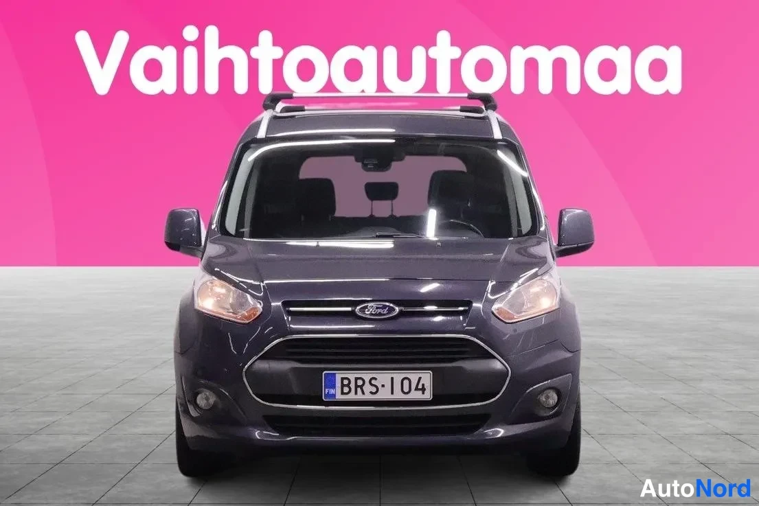 Edestä: Tummanharmaa Ford Tourneo edestäpäin, rekisterikilpi, kattokaiteet ja sumuvalot näkyvissä.
