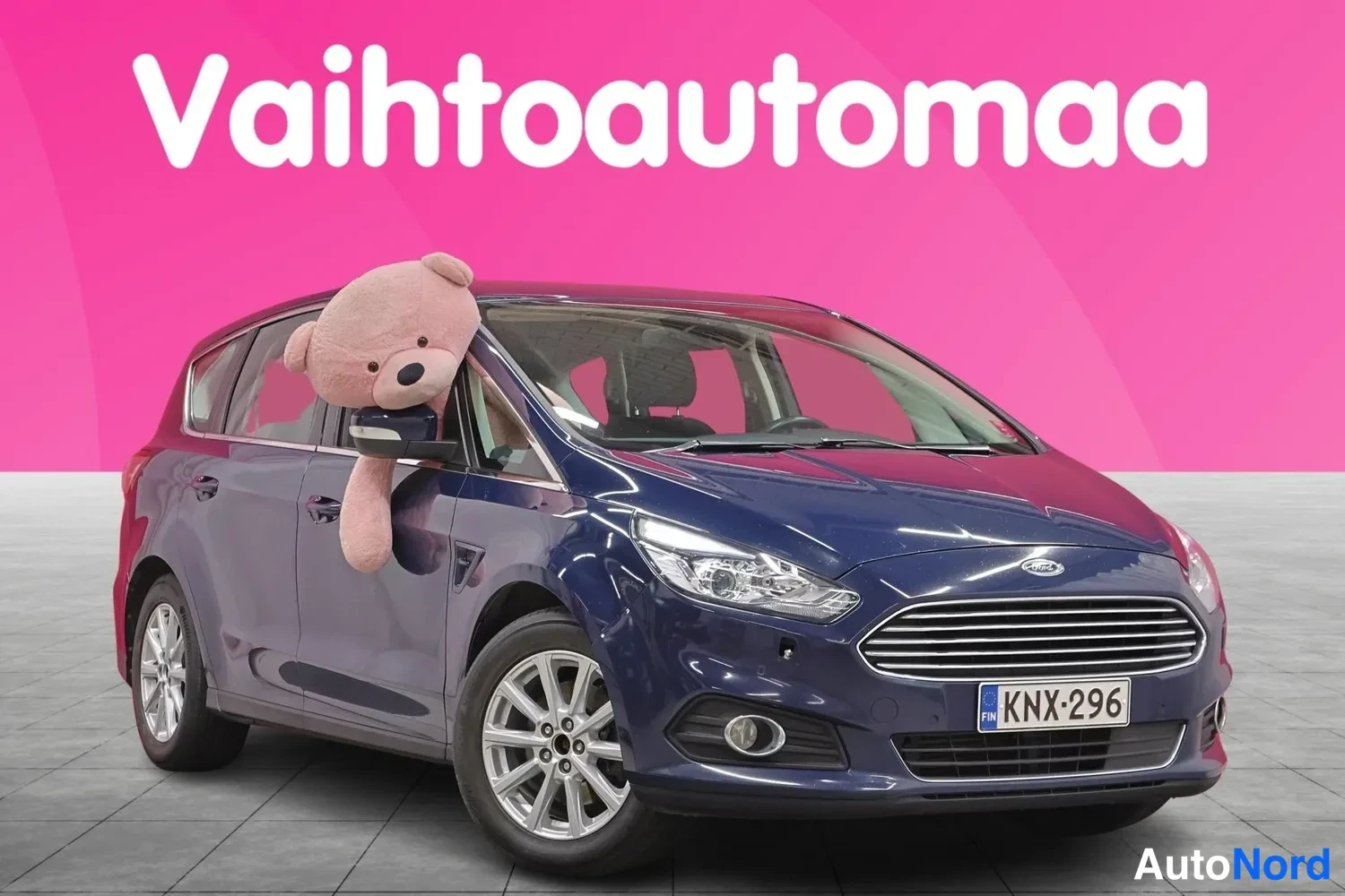 Etuviisto, nokka oikealle: Tummansininen farmariauto edestä oikealta kuvattuna, rekisterikilpi, suuri pehmolelu takanäytin peiliin kiinnitettynä.