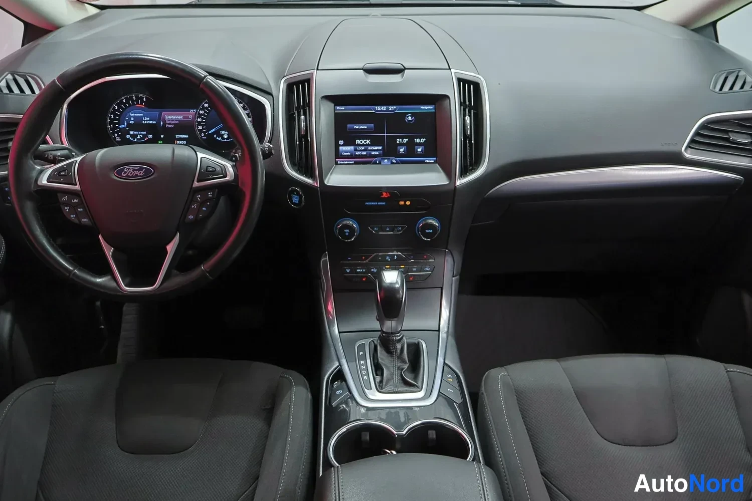 Yksityiskohta: Auton ohjaamo kuvattuna keskeltä: Ford-ohjauspyörä, mittaristo ja keskikonsolin infotainment-näyttö, jolla ROCK-soittolista ja lämpötilat 21,0° / 19,0°.