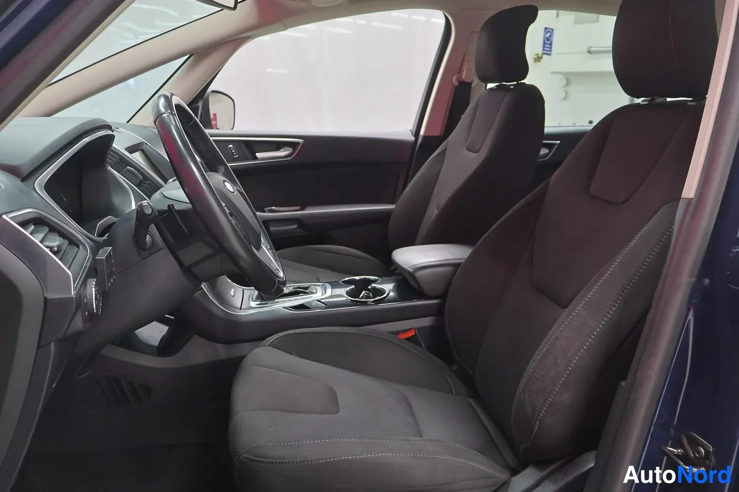 Muu: Sisätilakuva Ford S-MAX:stä: kuljettajan ja etumatkustajan tummat kangasistuimet, ohjauspyörä ja keskikonsoli kuppikeenkeillä näkyvät avatun ovelta.