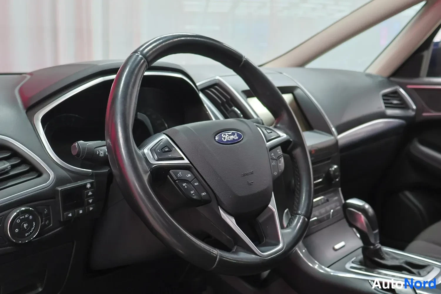 Muu: Ohjauspyörän lähikuva: Ford-merkki keskellä, monitoimipainikkeet ja automaattivaihteen valitsin kojelaudan takana.