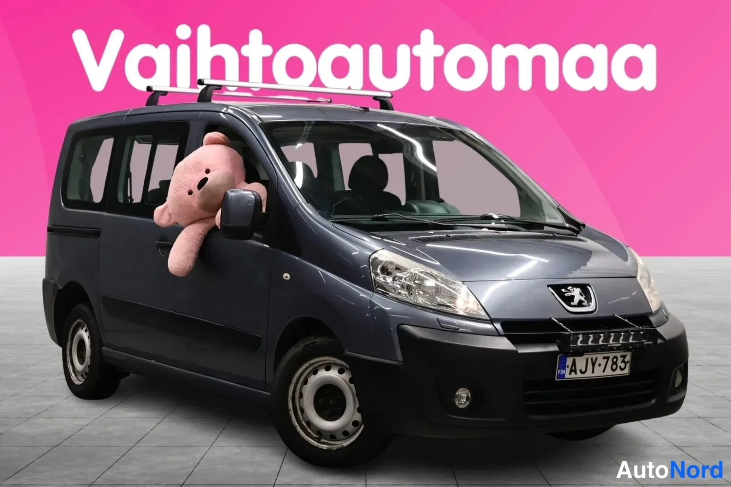 Etuviisto, nokka oikealle: Sinisenharmaa pakettiauto vinosti kuvattuna keula oikealla, kattotelineet ja pehmolelu ikkunassa.
