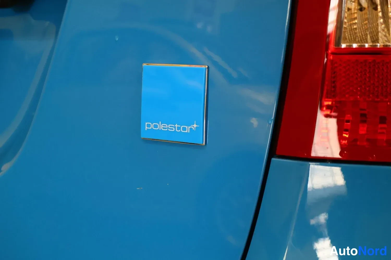 Yksityiskohta: Sininen Polestar-merkki autossa, taustalla punainen takavalo.