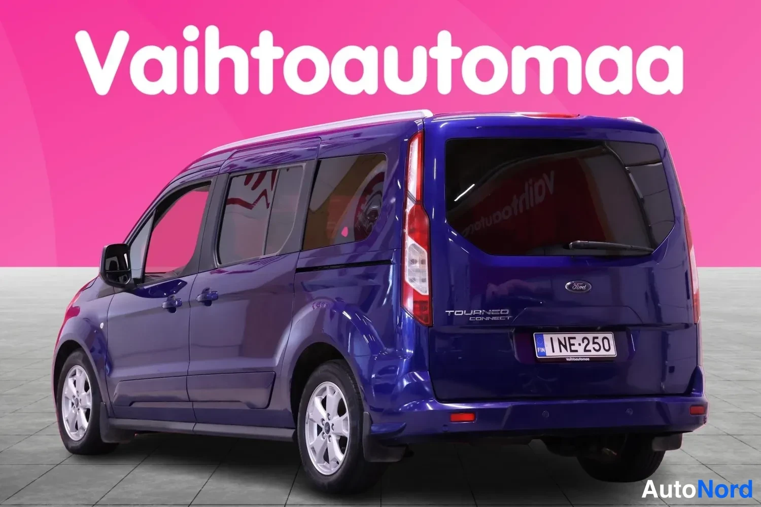 Takaviisto: Valkoinen Ford Tourneo Connect -pakettiauto vaaleanpunaisen taustan edessä. Takakulma näkyy, rekisterinumero ja takavalot. Auto on tummansininen.