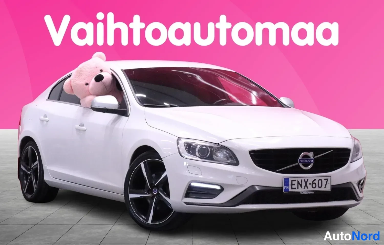Etuviisto, nokka vasemmalle: Kaksi etunäkymää: Valkoinen Volvo-sedani edestä ja kulmassa. Etusuojuksessa ruudut, kromattu Volvo-logo ja kyljellä mustat ja hopeiset vanteet.