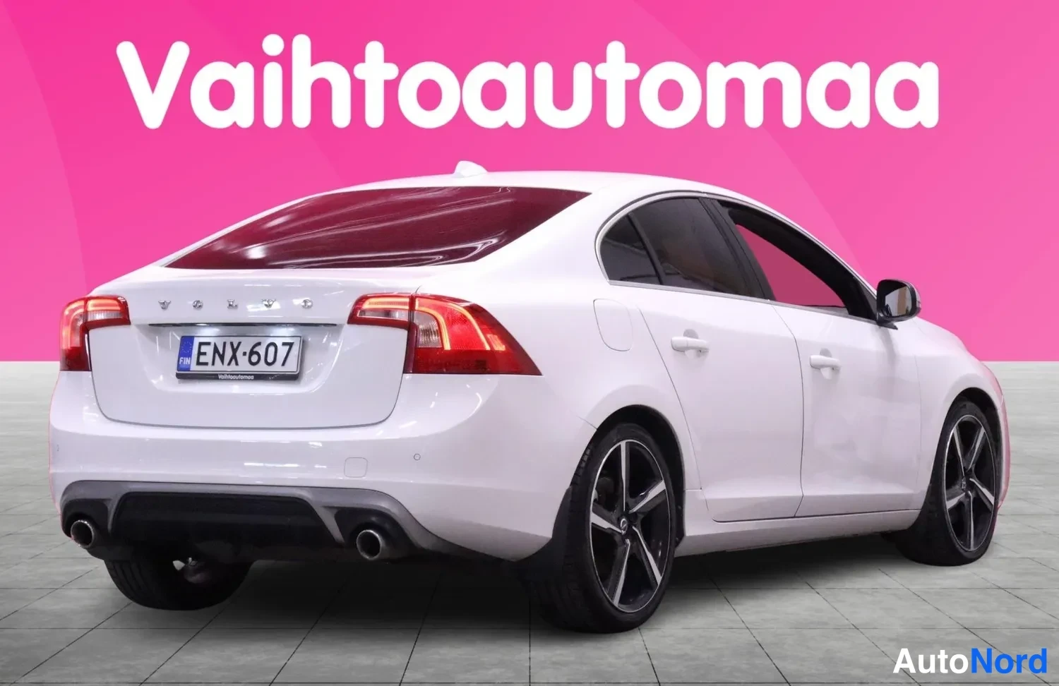 Takaa: Valkoinen Volvo-sedani takaa nähtynä. Takavalot, rekisterinumero, kaatokuorma-auto (matala maantie), takapuskurin alapuolella pakoputki. Teksti taustalla: Vaiht