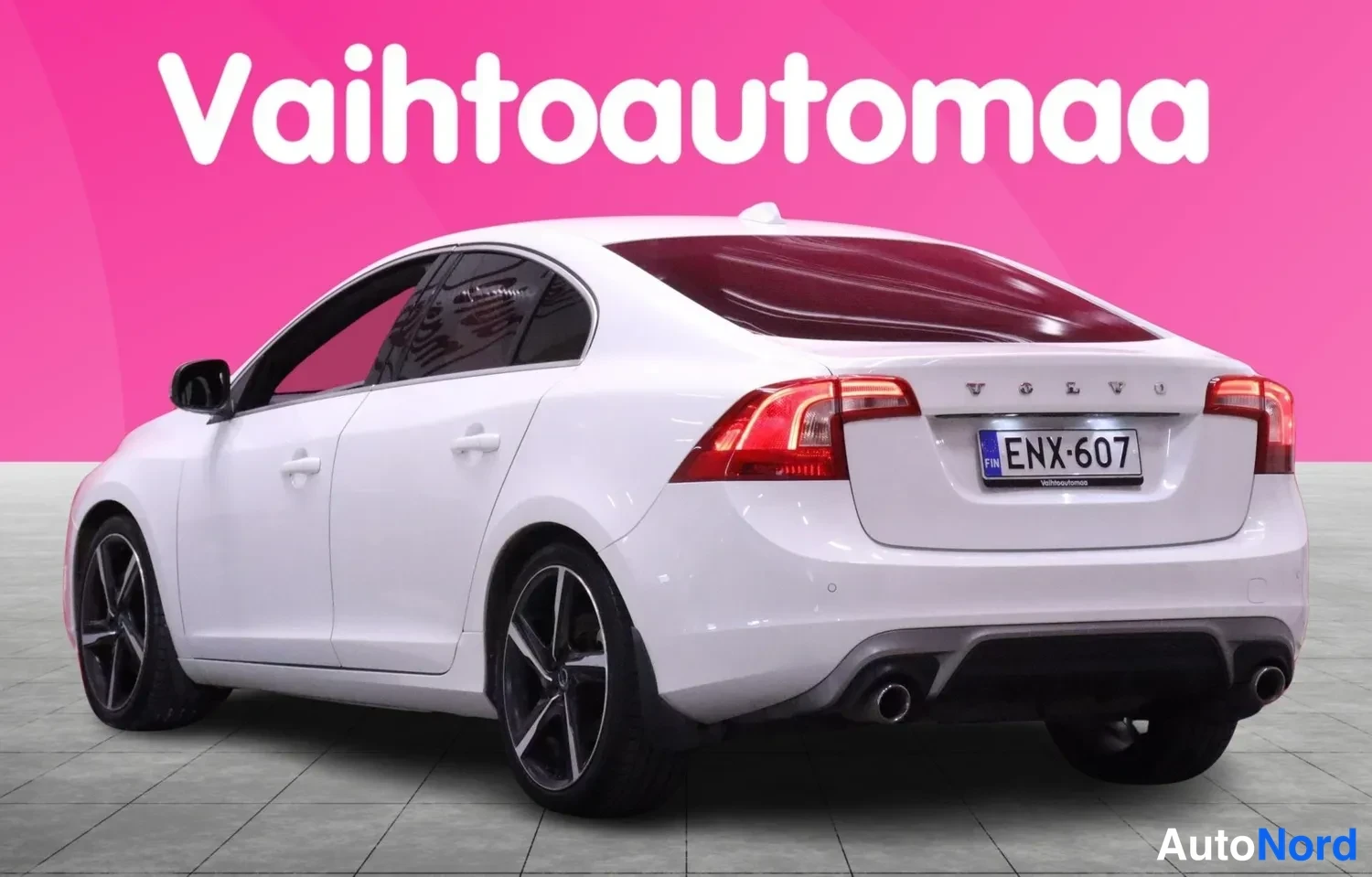 Takaviisto: Valkoinen sedan takaa ja vasemmalta sivulta viistosti kuvattuna. Takaluukissa VOLVO-kirjaimet, rekisterikilpi, kaksi pakoputkea. Taustalla vaaleanpunainen Vaiht