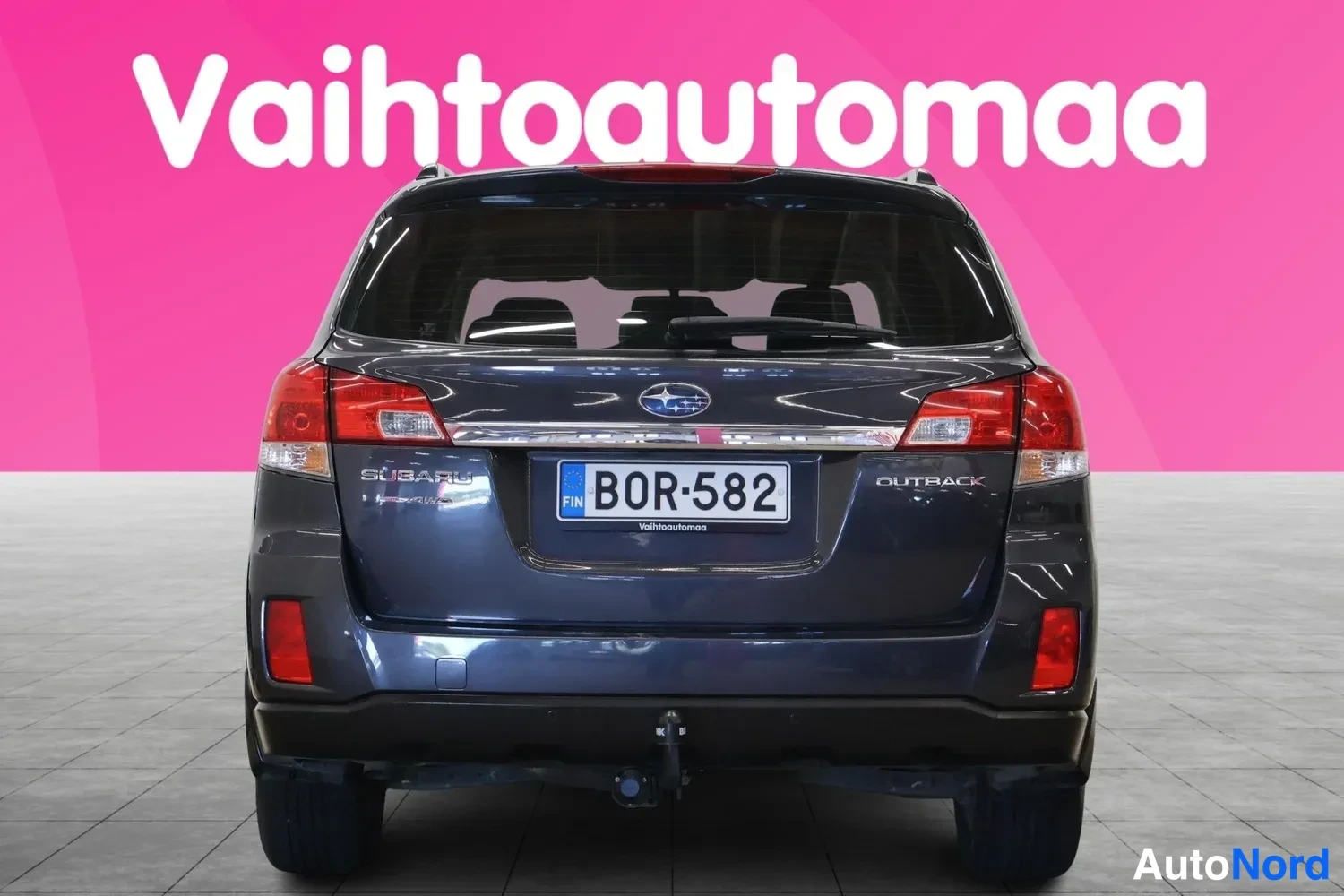 Takaa: Suora taka­kuva: tummanharmaan farmariauton takavalot, Subaru-merkki, -kilpi ja vetokoukku.