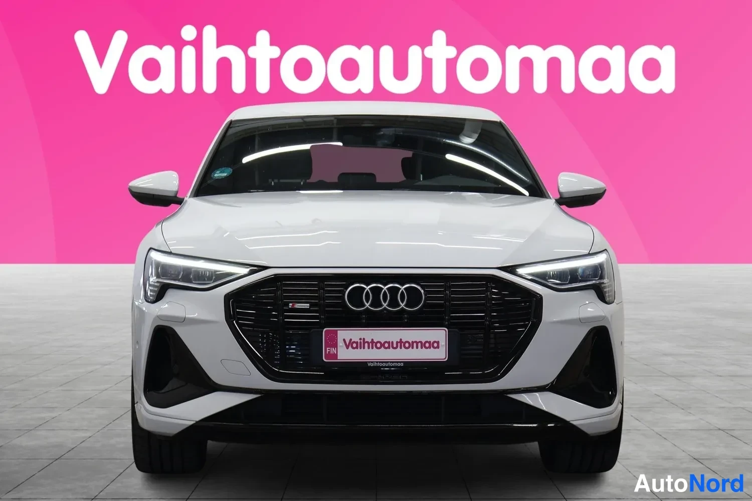 Edestä: Valkoisen Audin etunäkymä, musta etusäleikkö ja Audi-merkki, Vahtoautomaan mainoskyltti rekisterikilven paikalla