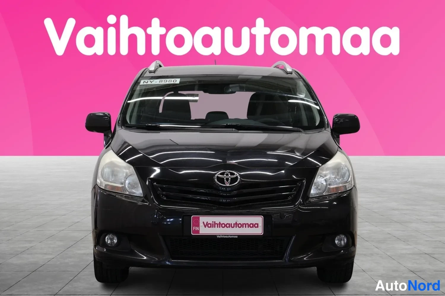 Edestä: Musta Toyota Verso -henkilöauto suoraan edestä,Toyota-merkki ja etupuskuri näkyvissä.