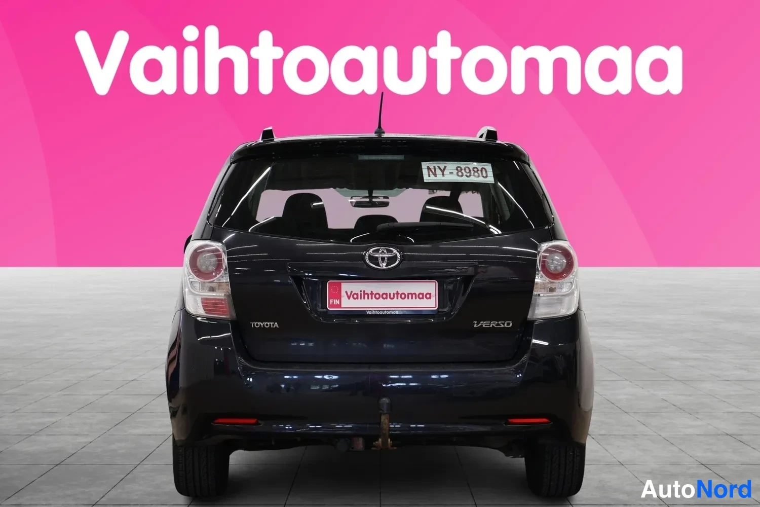 Takaa: Auton takaosa suoraan takaa: musta Toyota Verso, rekisterikilpi NY-8980, takavalot ja vetokoukku näkyvissä, taustalla vaaleanpunainen Vaihtoautomaa-tausta.