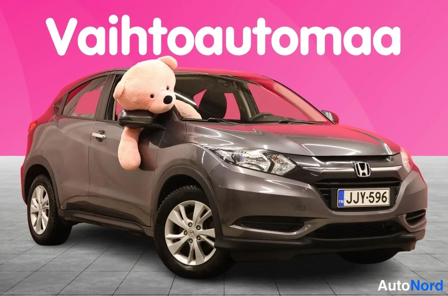 Etuviisto, nokka oikealle: Front three-quarter view of dark grey Honda HR-V, license plate, teddy bear leaning out of driver's side window.