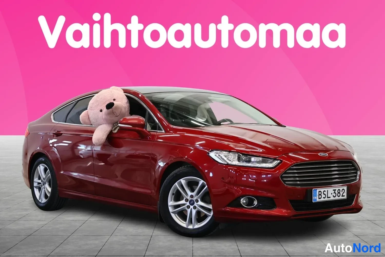 Etuviisto, nokka oikealle: Punainen Ford Mondeo -henkilöauto etu-VAS-kulmasta, etupuoli ja kylki näkyvät; suurennettu nallekeinonukke pökertää kuljettajan ikkunasta ulos.