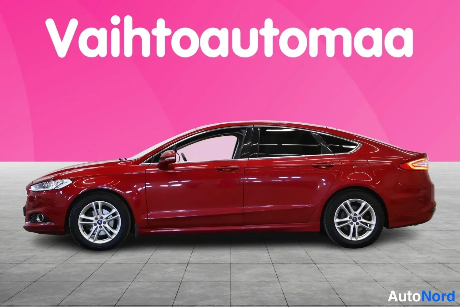 Sivulta: Punainen Ford Mondeo näkyy sivusta kirkkaanpunaisen taustan edessä. Auto seisoo harmaalla laatoitetulla lattialla. Hopeiset kevytmetallivanteet ovat selvästi nä