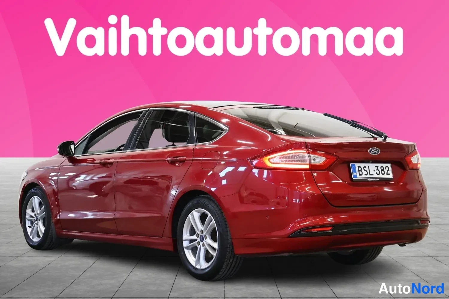 Takaviisto: Punainen Ford Mondeo takasivasta kuvattuna kirkkaanpunaisen 'Vaihtoautoma' tekstin edessä. Takaluukussa on Ford logo ja rekisterikilpi.